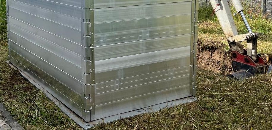 Großer Metallcontainer auf Freigelände mit Bagger daneben.
